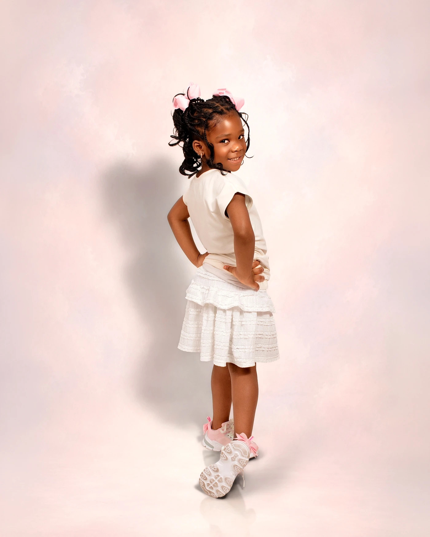 A child in white looking over her shoulder against a pink studio backdrop