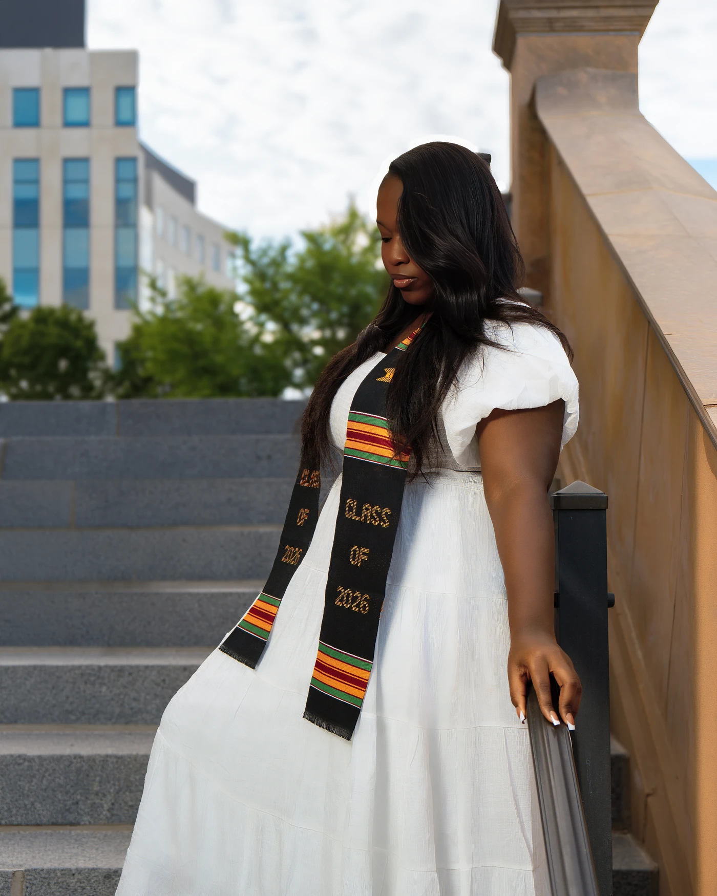 A graduate in a white dress and graduation stole on outdoor stairs