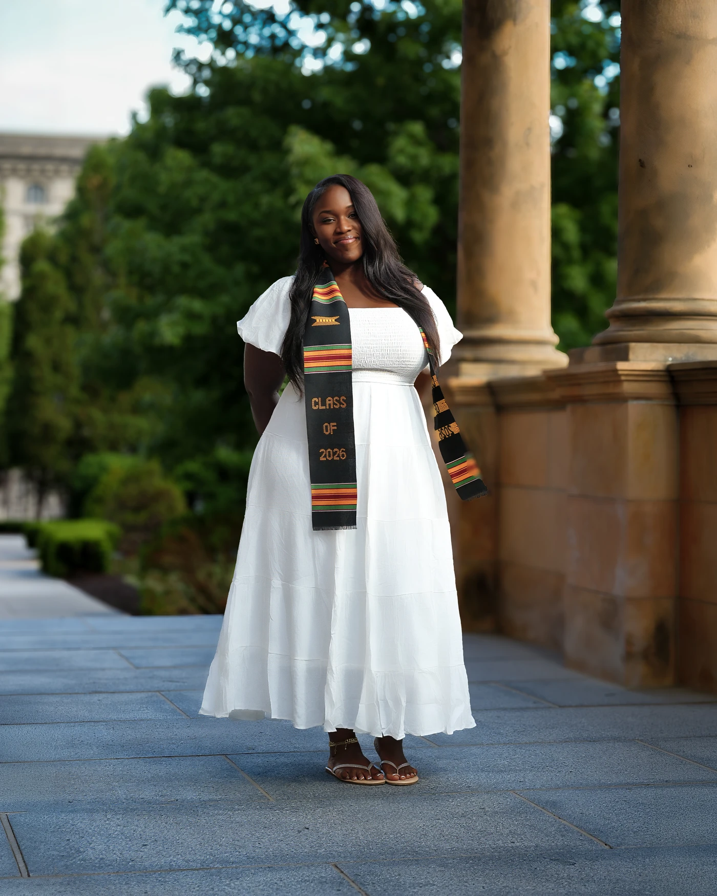 A smiling graduate in a white dress and graduation stole beside outdoor columns