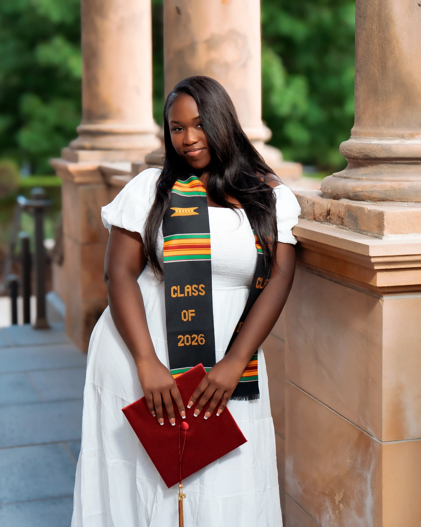 A graduate holding a red mortarboard beside stone columns
