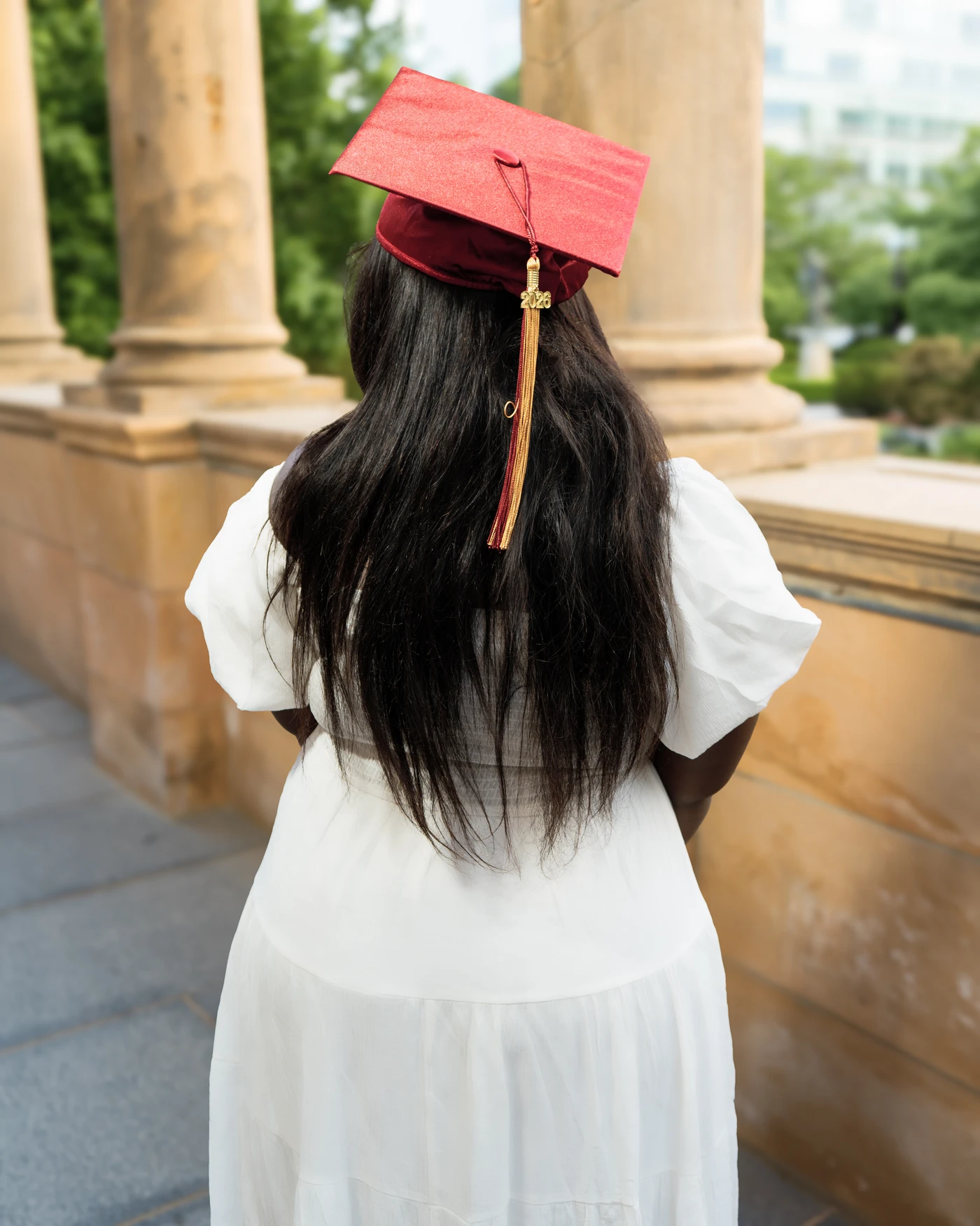 A graduate wearing a red mortarboard beside outdoor columns