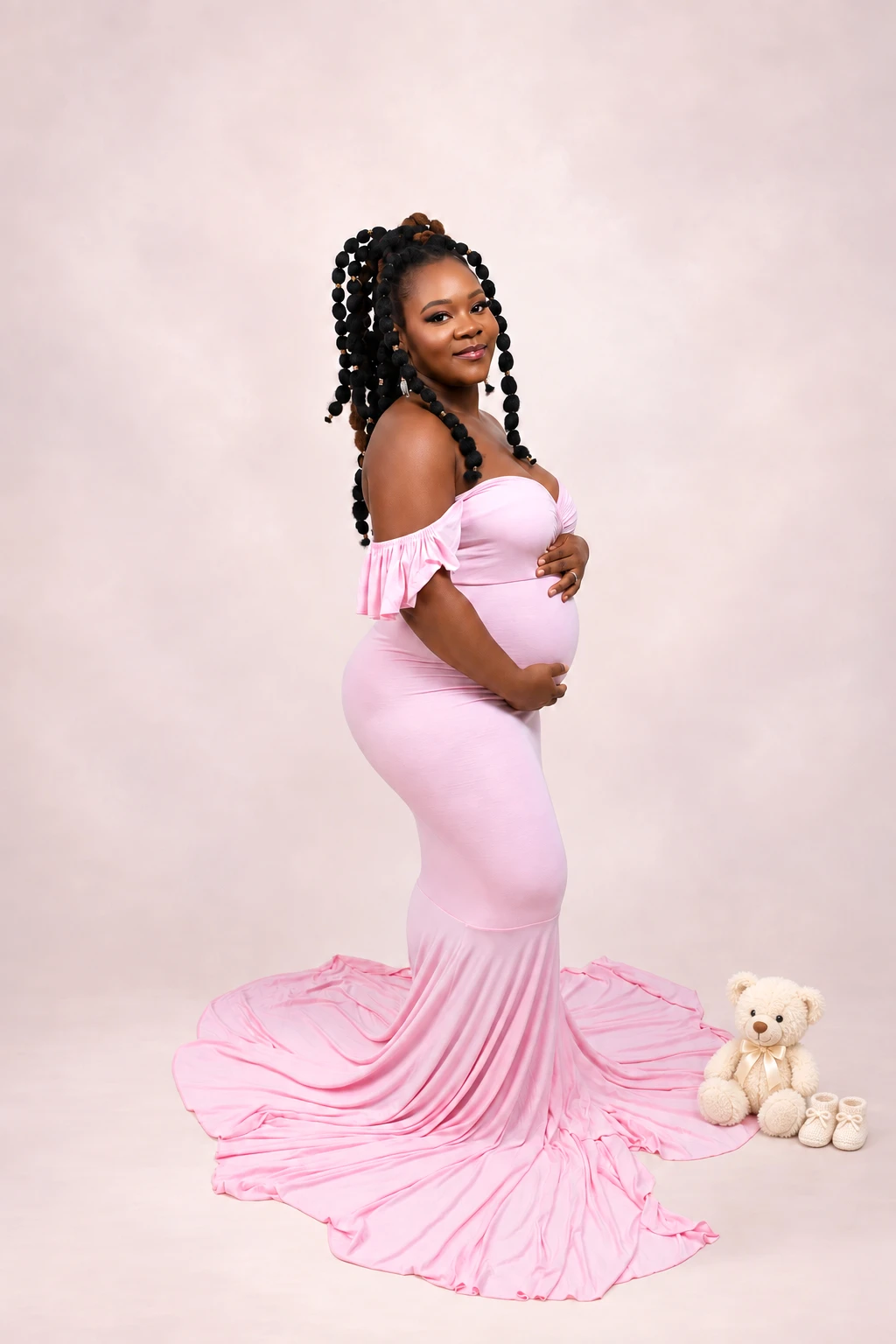 A mother-to-be in a pink gown beside a teddy bear and baby shoes