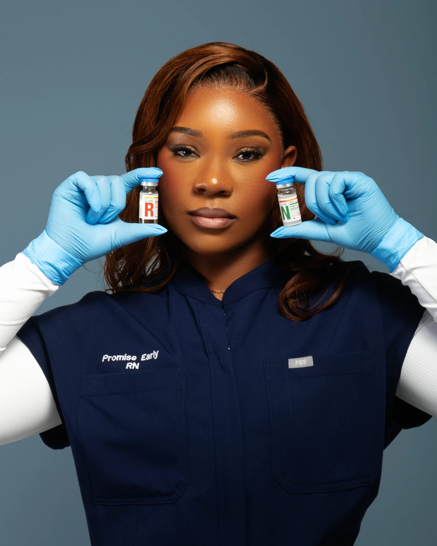 A close-up portrait in navy scrubs and blue gloves holding two small vials