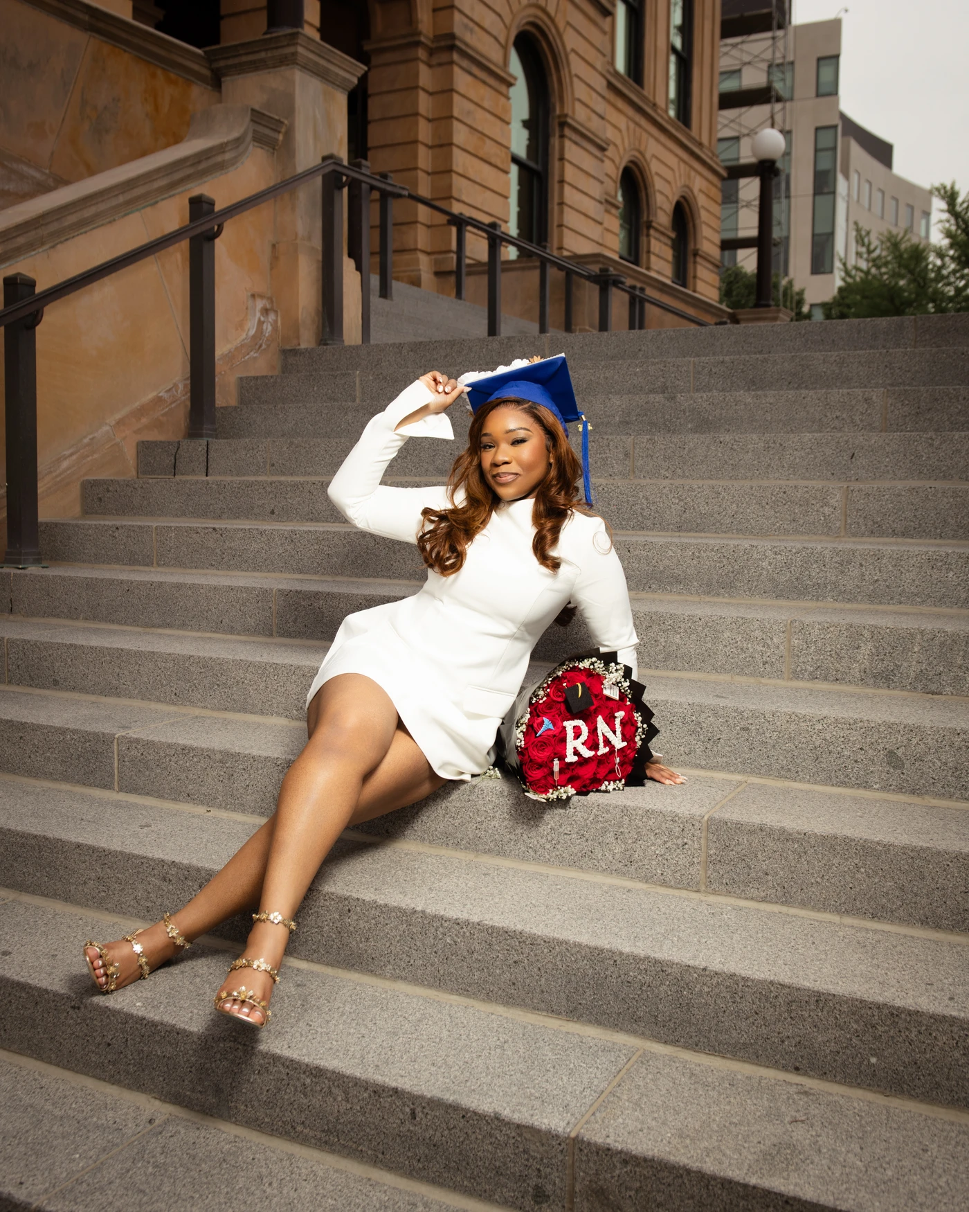 A graduate in a blue mortarboard seated on outdoor stairs beside a rose bouquet